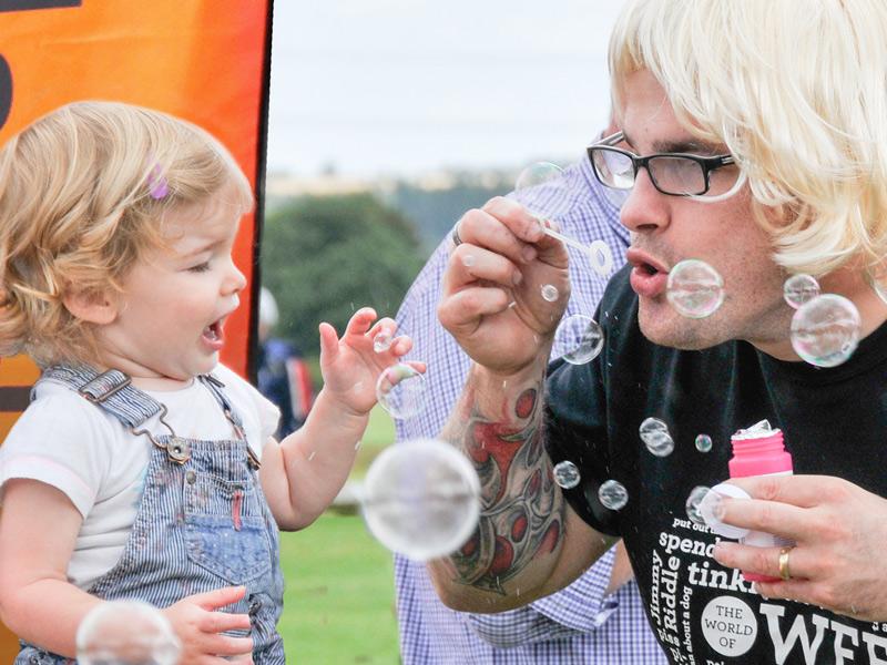 Man in a blonde wig blows bubbles with a small child who is delighted!